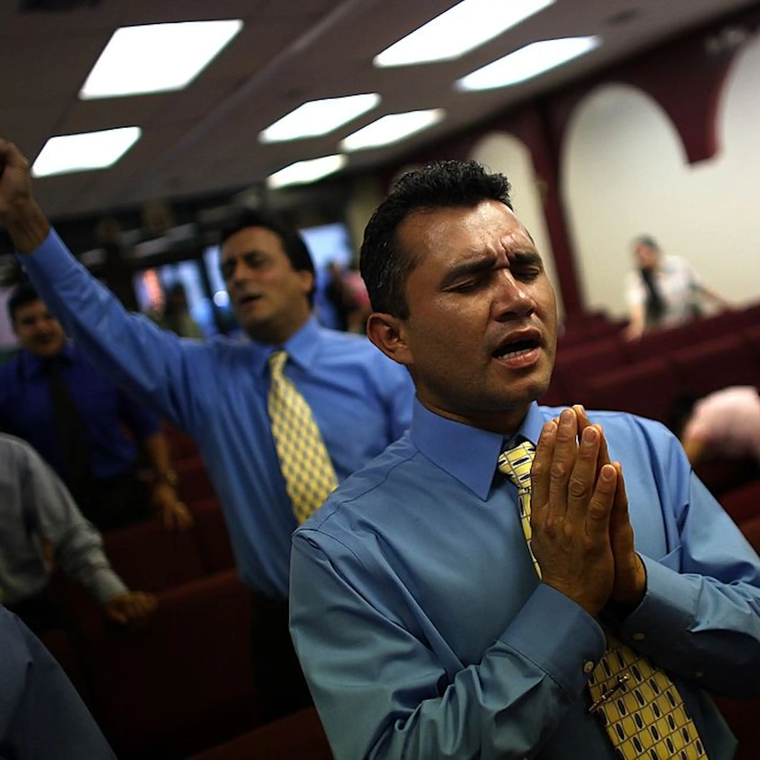 Two men wearing ties and blue button-up shirts, with their eyes closed in prayer, stand inside a room with rows of red seats.