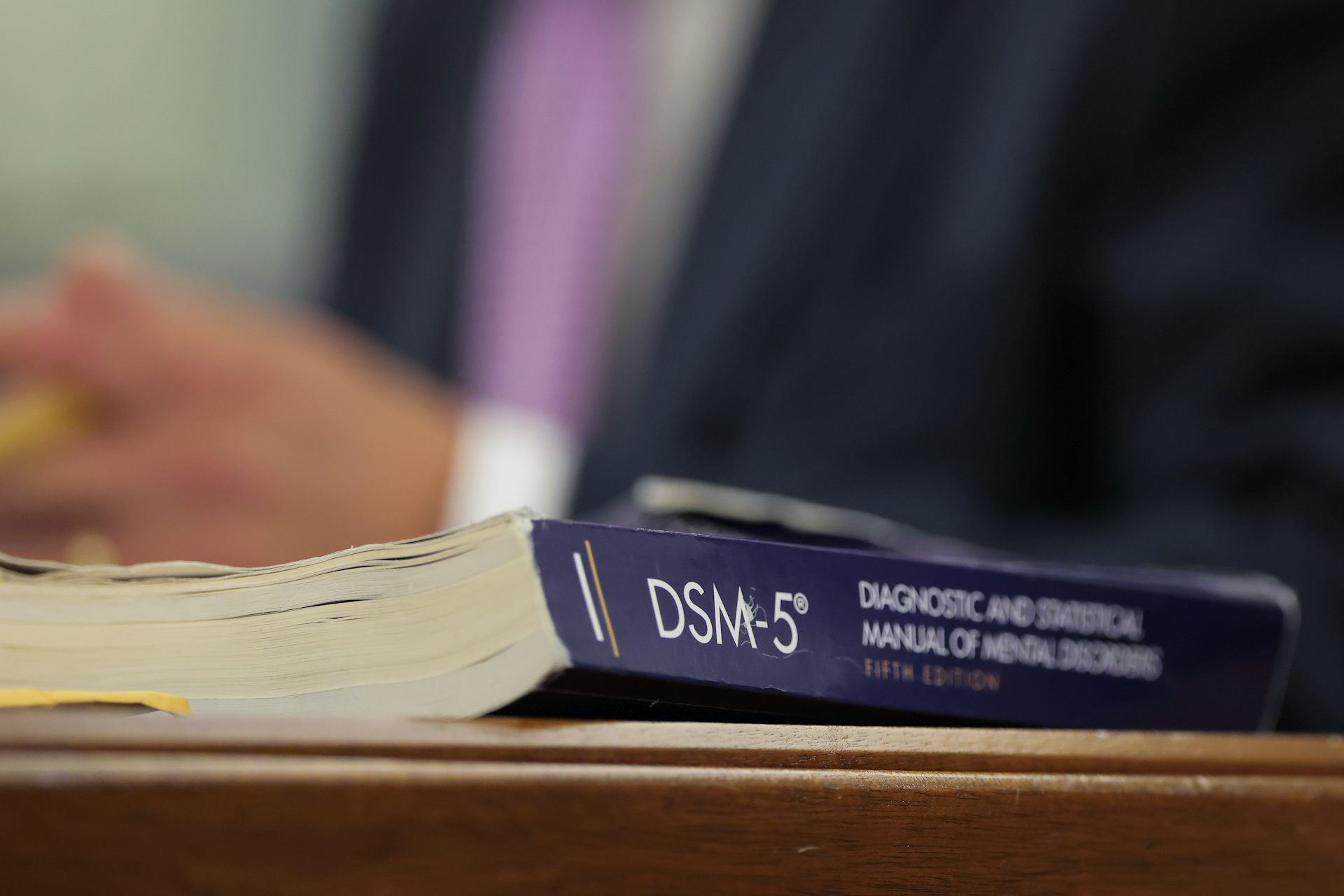 A book on table or desk titled DSM-5 with blurred torso of man in suit with hands clasped in background