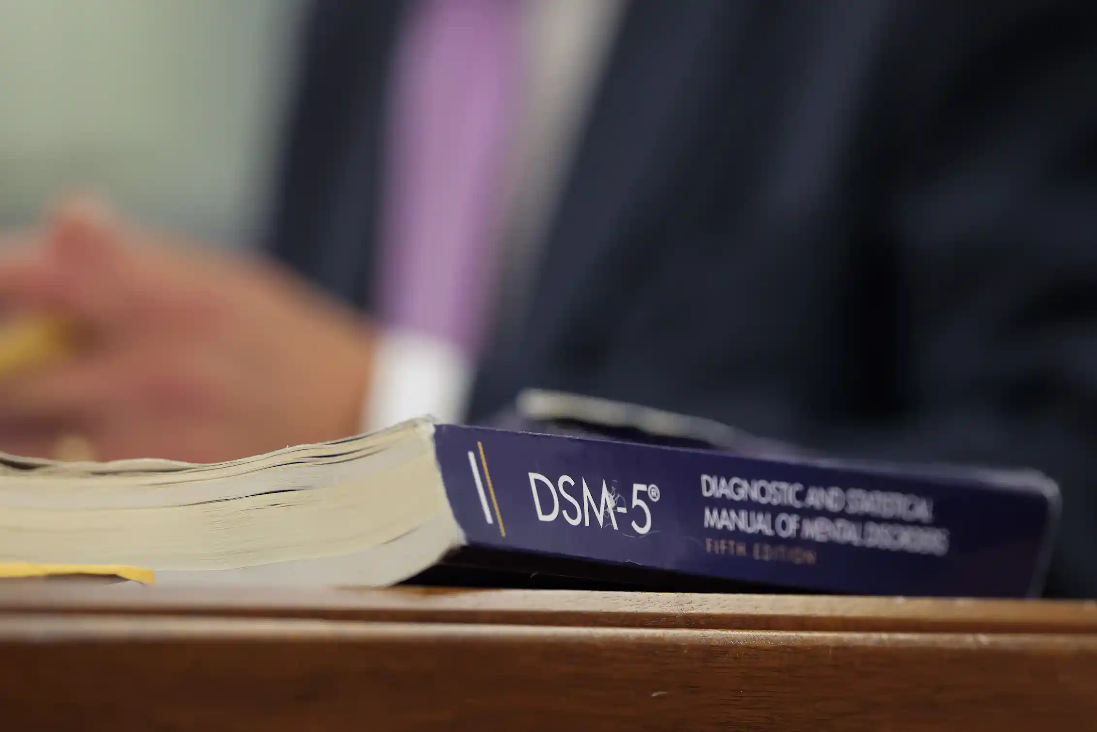 A book on table or desk titled DSM-5 with blurred torso of man in suit with hands clasped in background