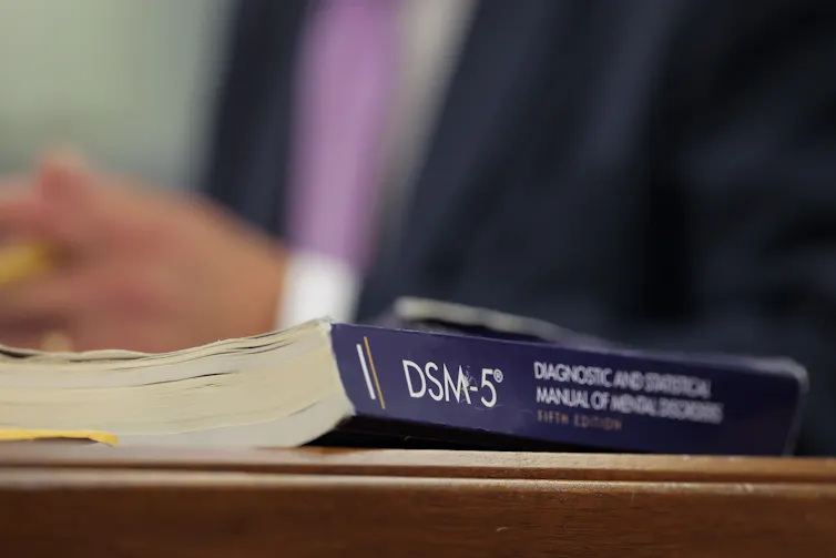 A book on table or desk titled DSM-5 with blurred torso of man in suit with hands clasped in background
