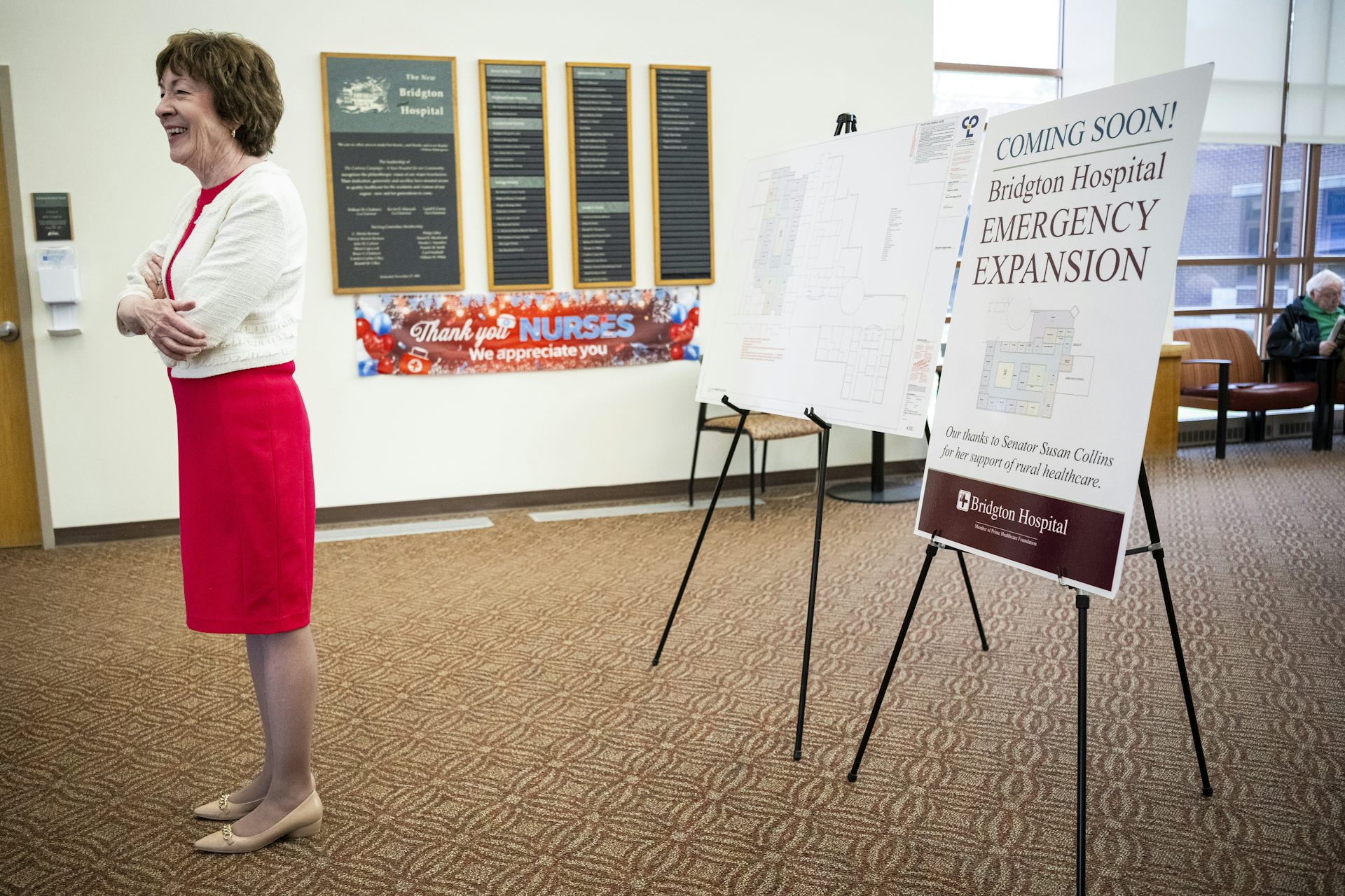 A woman with dark hair in a red dress and white sweater, standing in a large room with posters behind her.