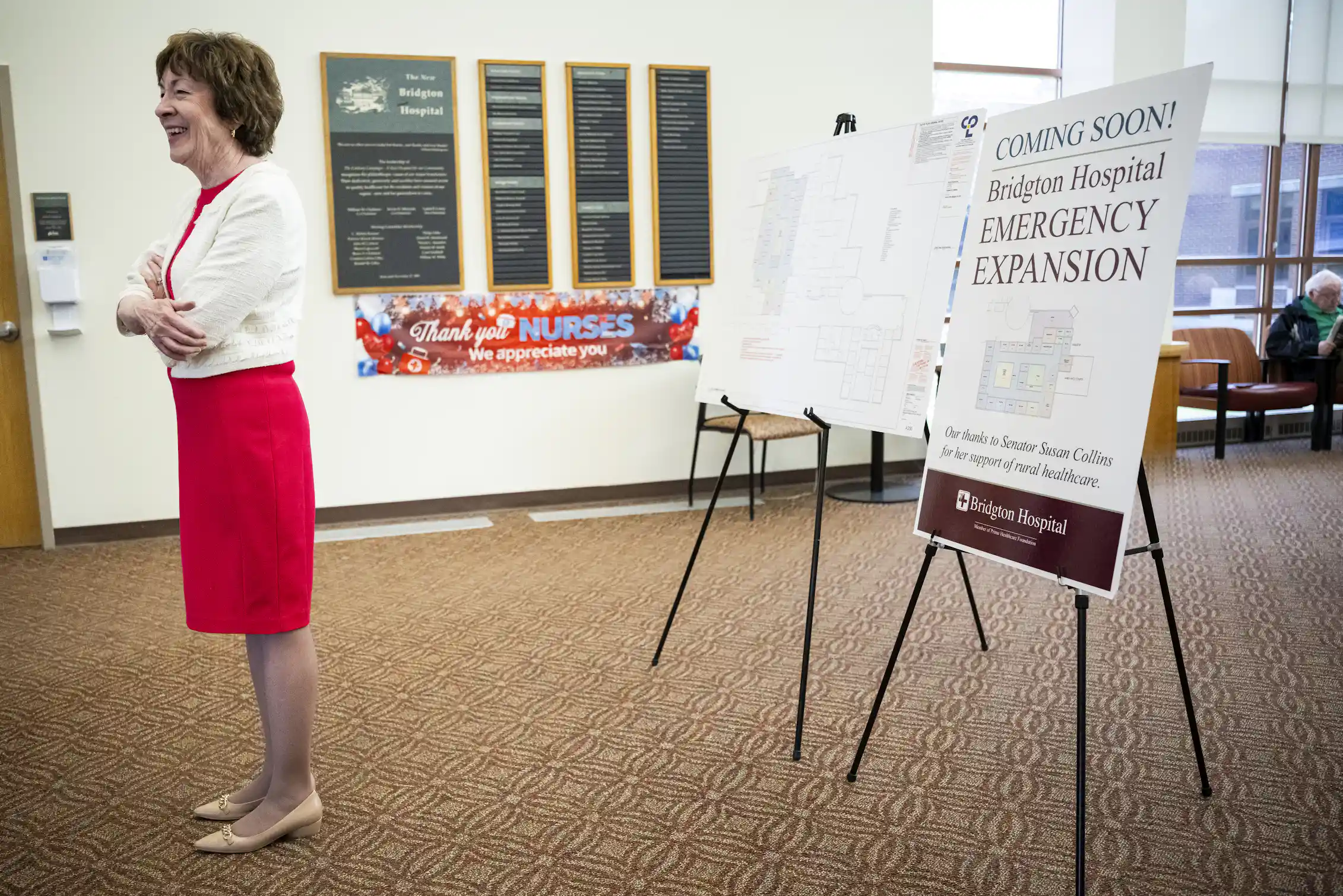 A woman with dark hair in a red dress and white sweater, standing in a large room with posters behind her.