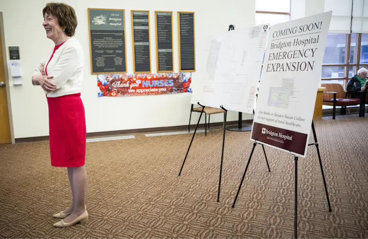 A woman with dark hair in a red dress and white sweater, standing in a large room with posters behind her.