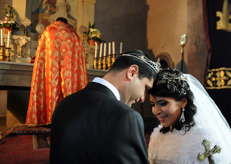 A couple wearing silver crowns press their foreheads together and smile as a priest in an orange and gold robe stands nearby.
