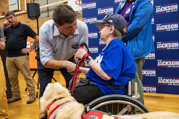 A dark-haired man in a blue shirt greets a person in a wheelchair holding a dog on a leash.