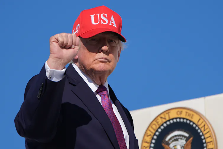 Trump boards Air Force One, holds up his fist, wearing a red cap that says USA.