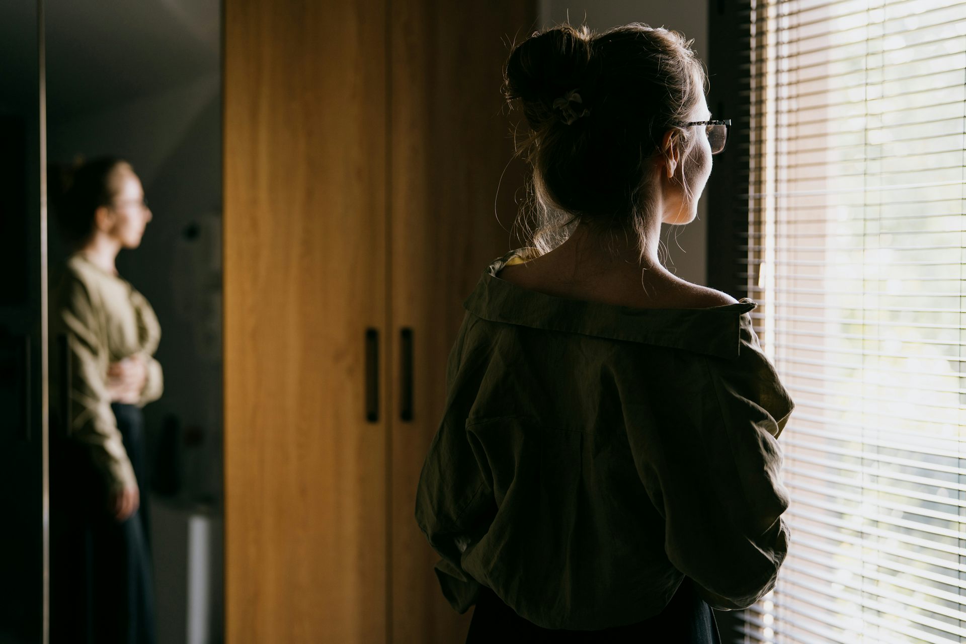A young woman pictured from the back gazing out a window with open blinds.