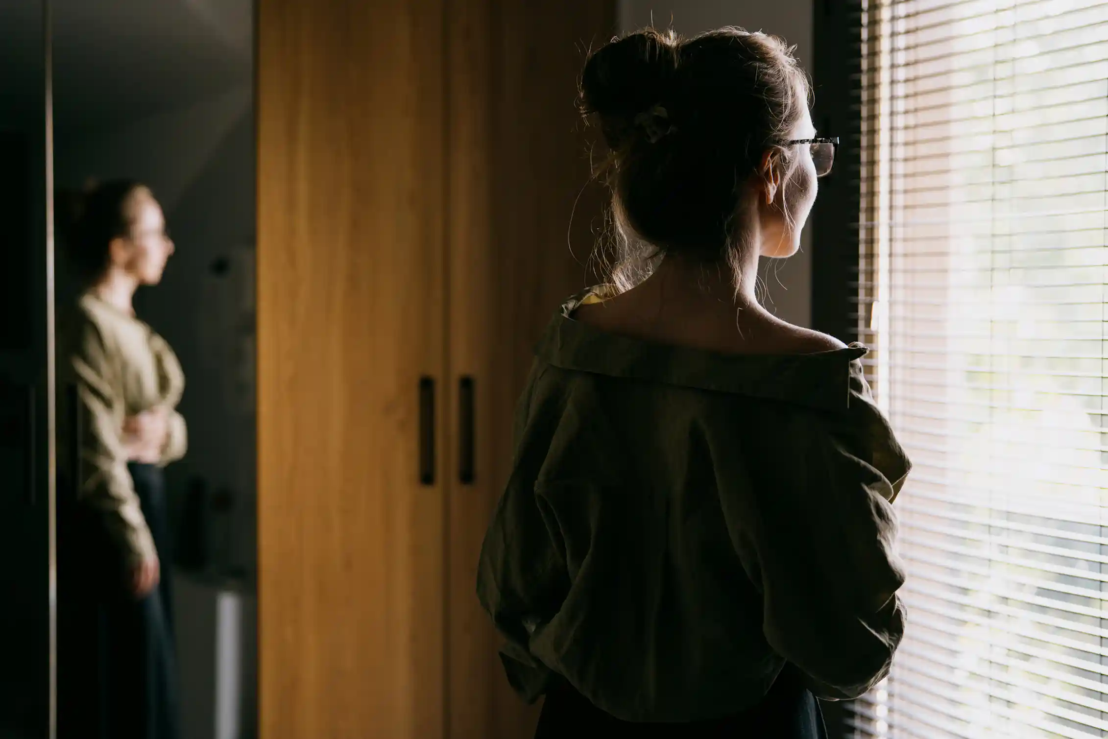 A young woman pictured from the back gazing out a window with open blinds.