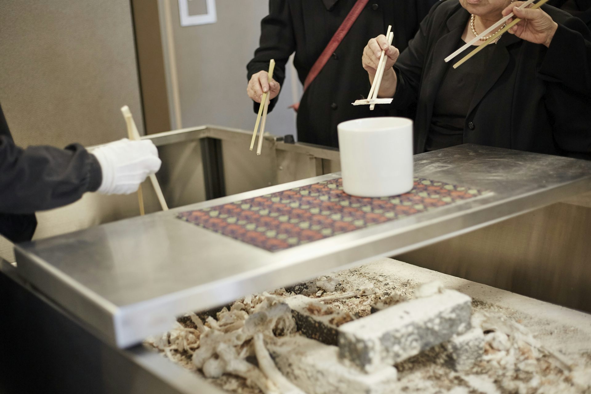 People dressed in black, holding large chopsticks, putting bone fragments in a funerary urn.