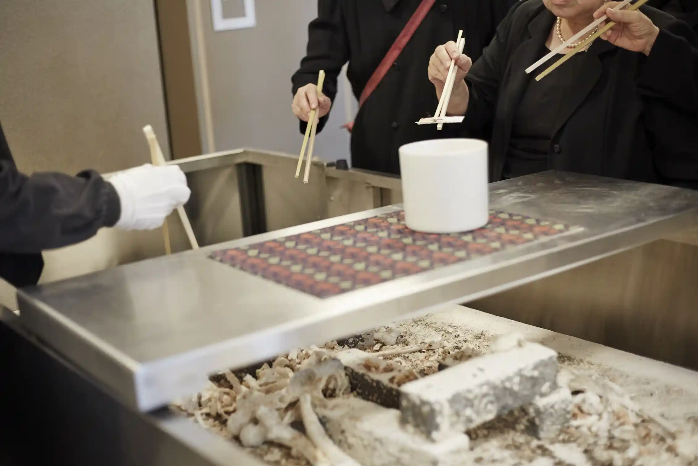 People dressed in black, holding large chopsticks, putting bone fragments in a funerary urn.