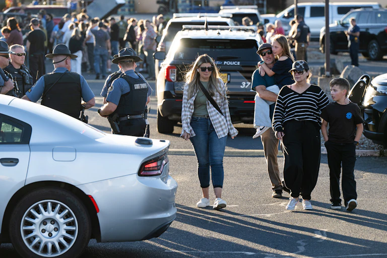 A man carries a child as he and others walk past law enforcement officers and police vehicles in a crowded parking lot
