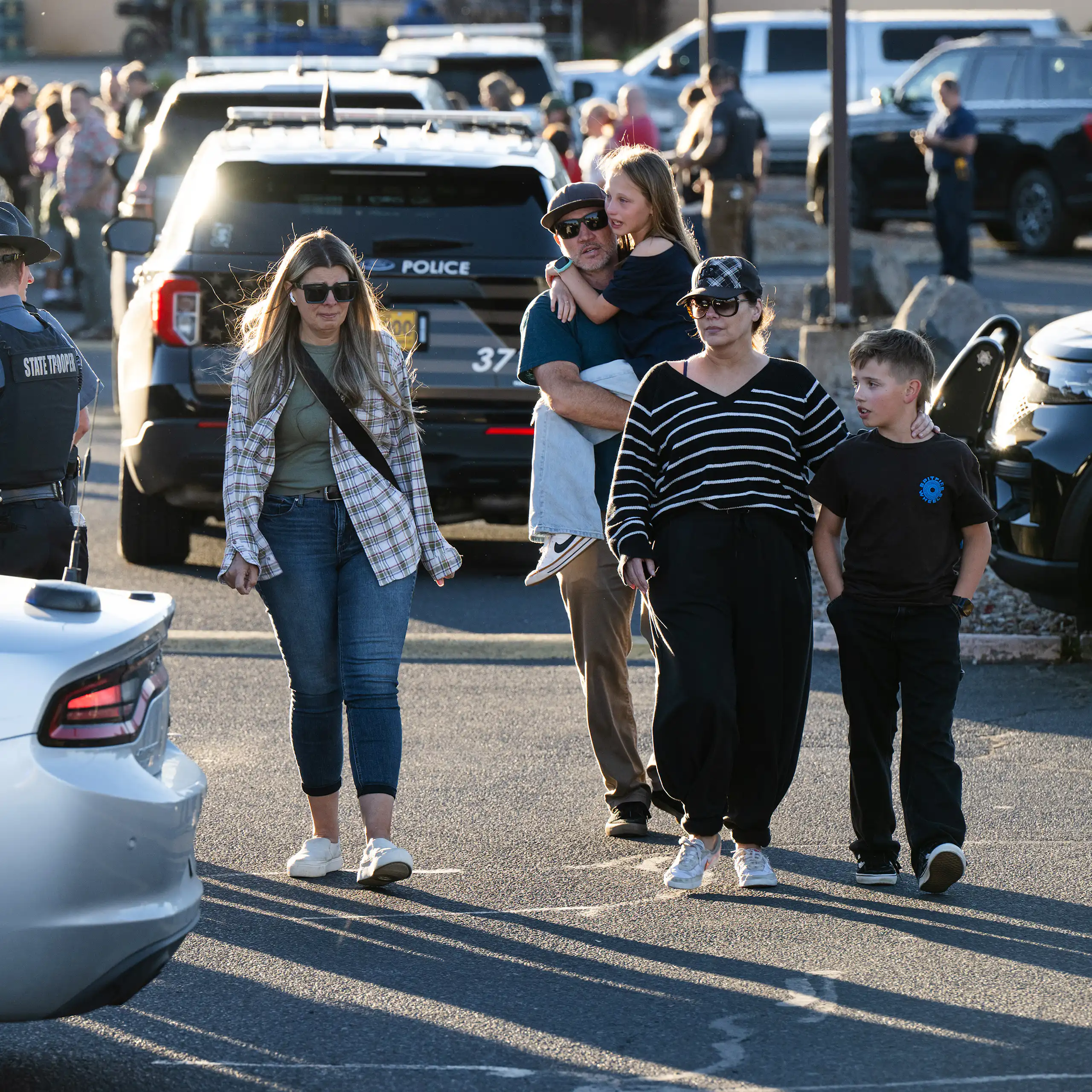 A man carries a child as he and others walk past law enforcement officers and police vehicles in a crowded parking lot