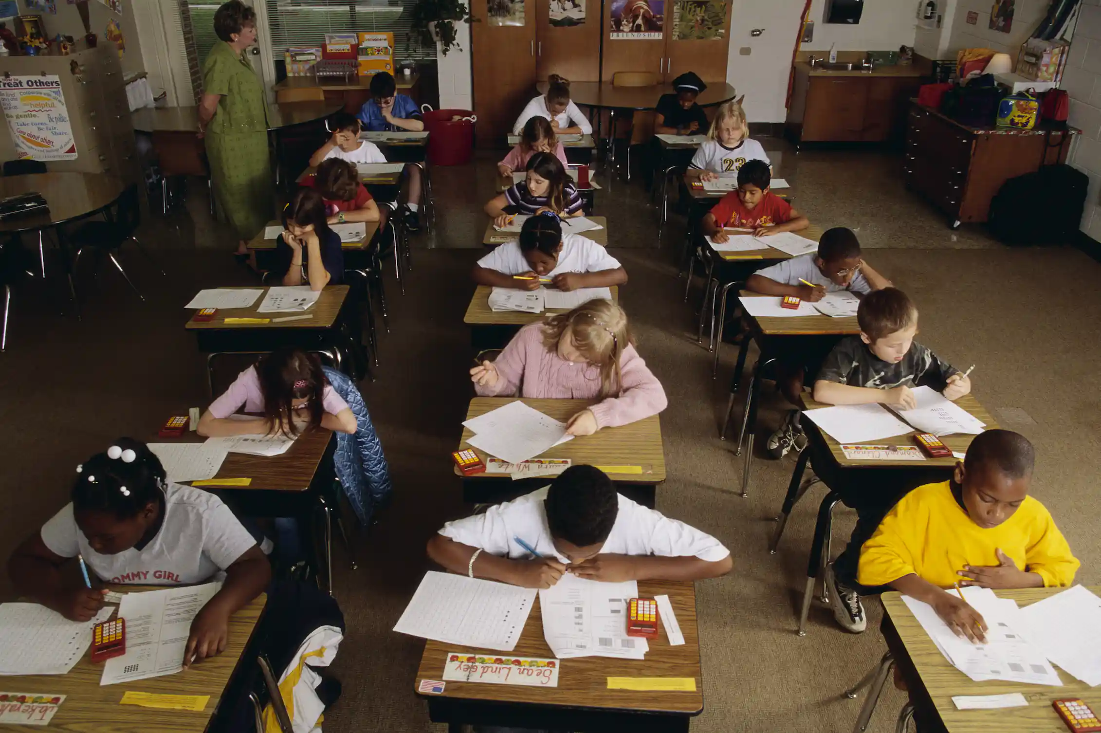 Three rows of children sit at desks and lean over white test books.