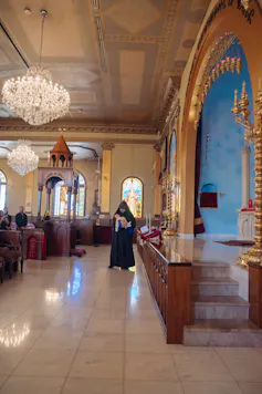 A priest in black robes and a black hood walks through a sanctuary, holding a baby.