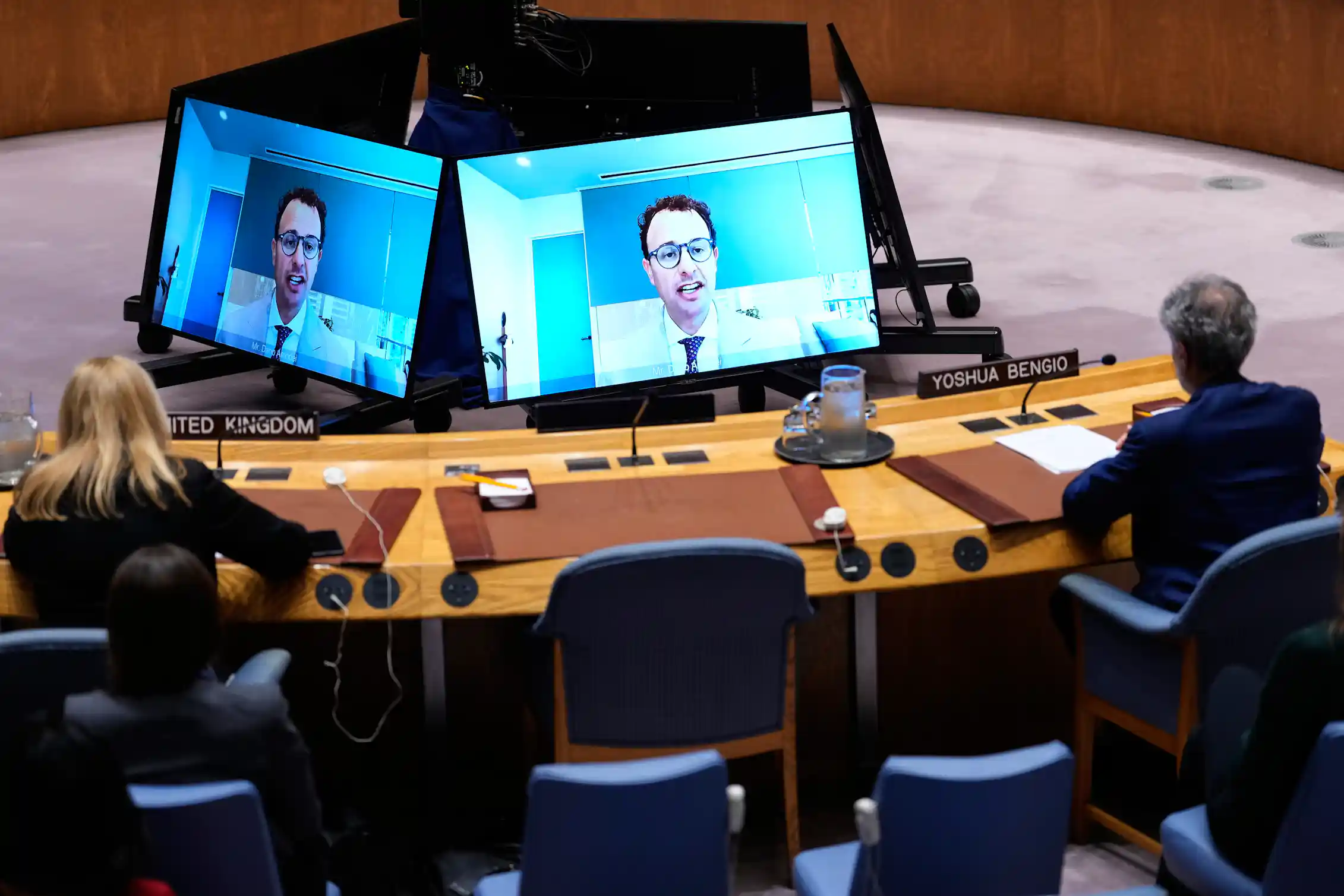 Man wearing tie and classes appears on video monitors in front of people seated at long table