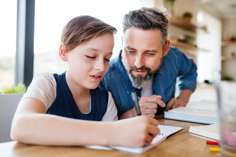 A father and a son at a table and the father is watching what the son writes.