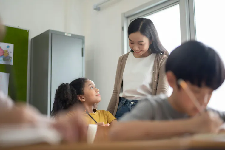 A teacher smiles in conversation with a preteen student who is seated at a desk.