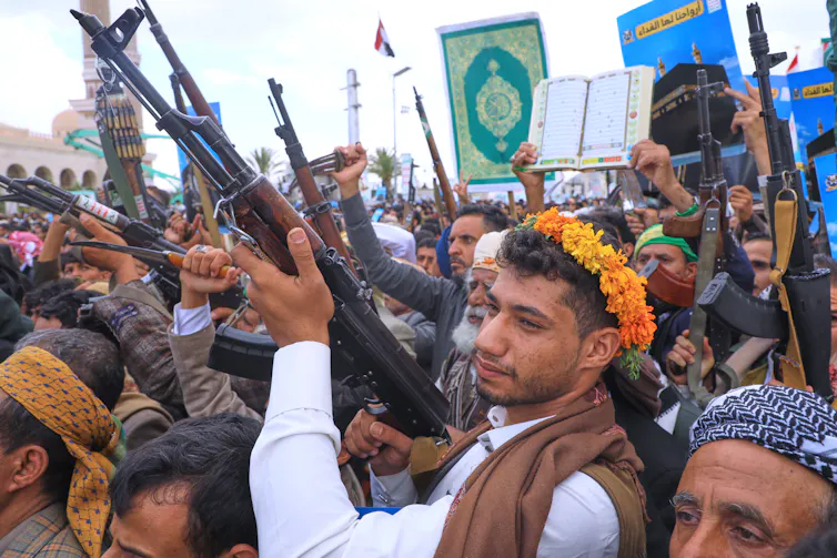 Young men in a crowd raise machine guns.