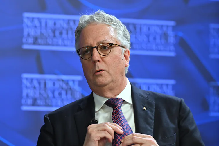 A photograph of Mark Scott, in a suit and tie, in front of a blue backdrop that reads 'National Press Club'.