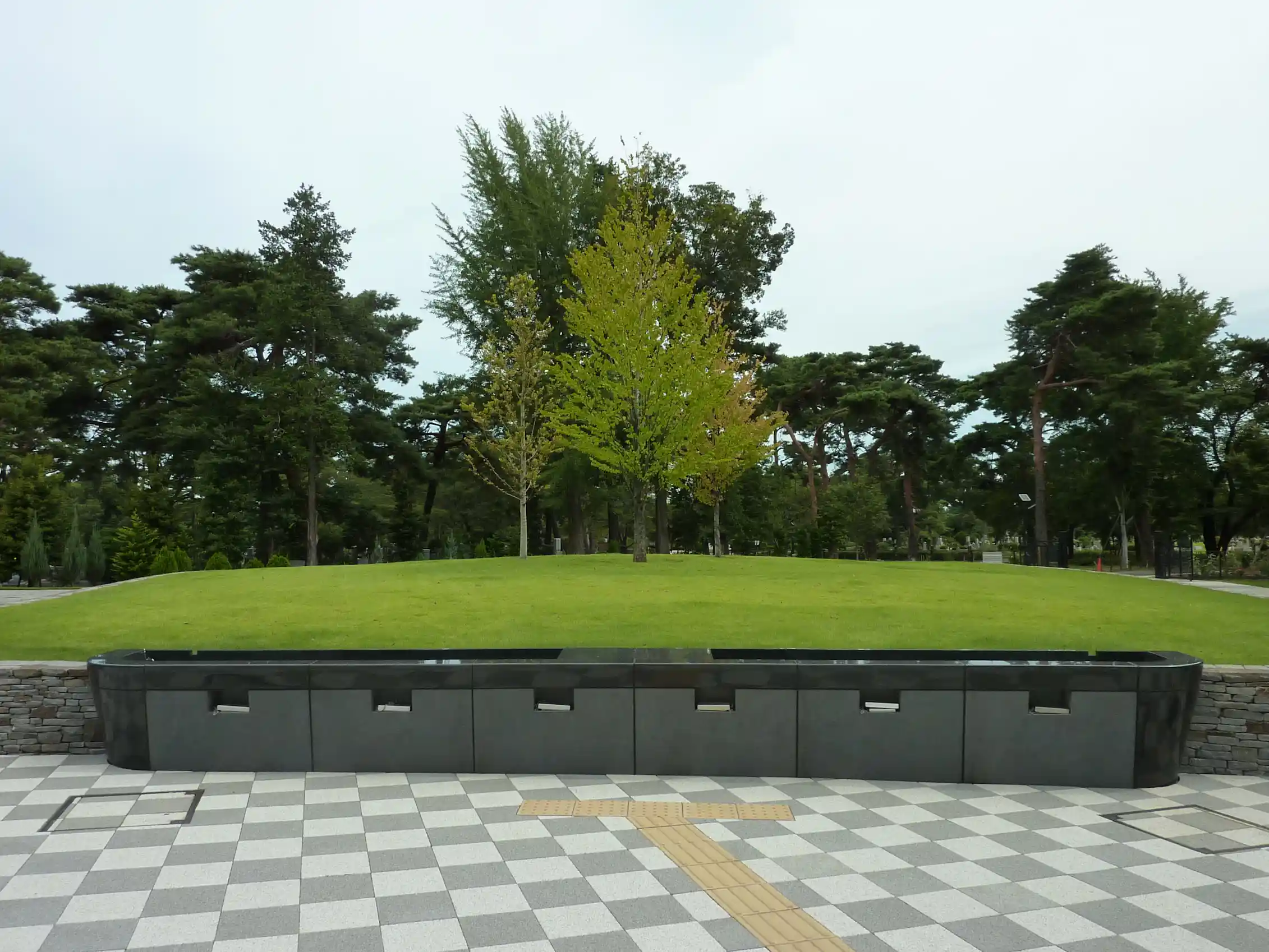 A row of dark stone compartments in front of a grassy mound and trees.