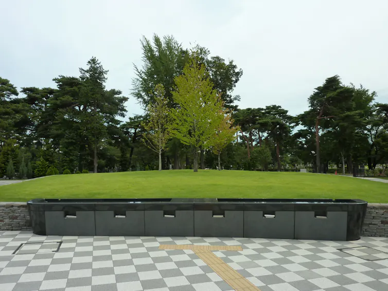 A row of dark stone compartments in front of a grassy mound and trees.