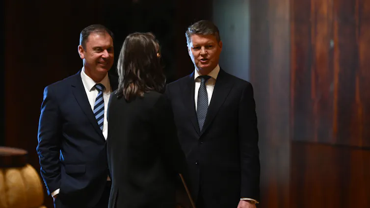 Two men wearing suits speak to a third person who is facing away from the camera.