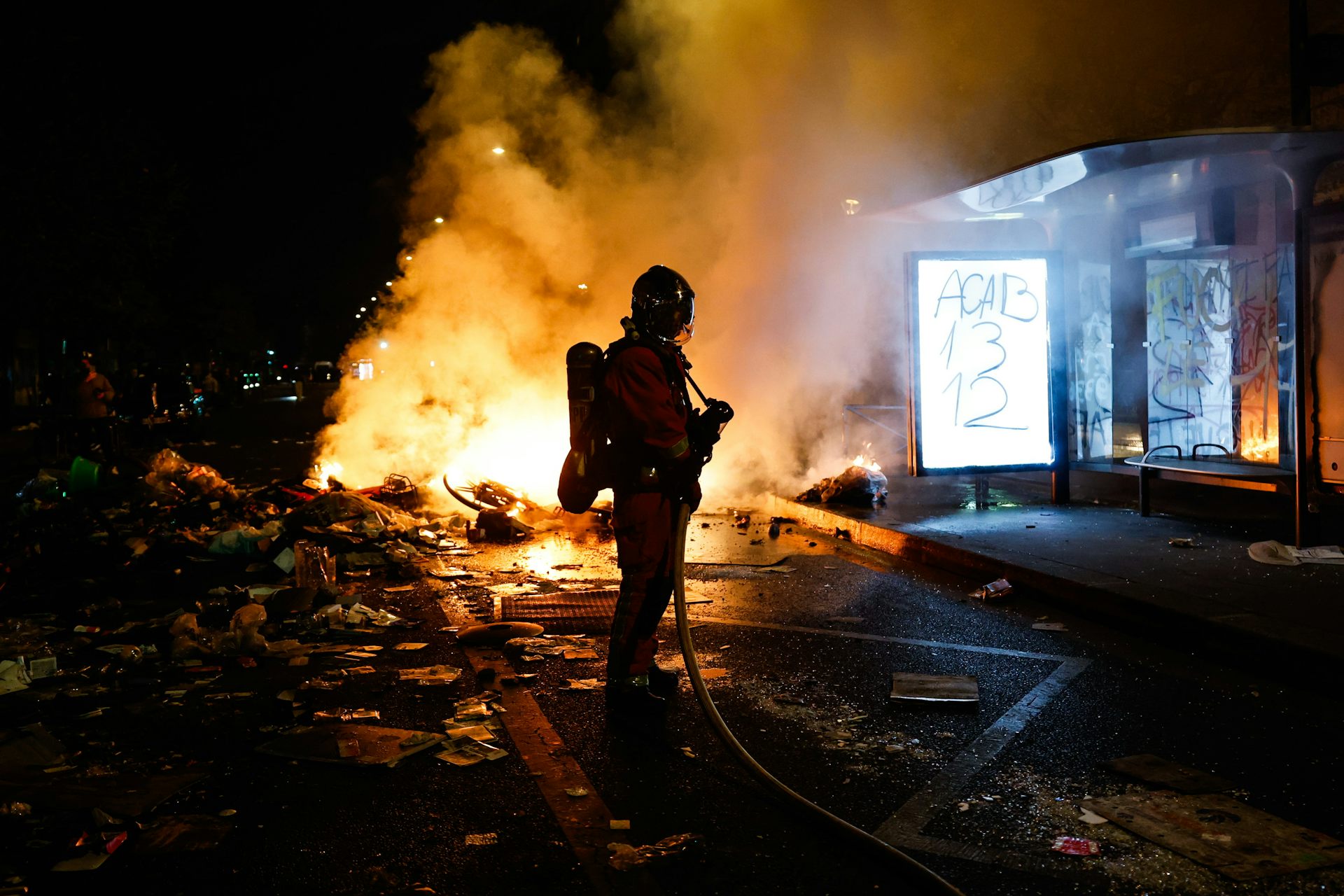 Un bombero en la calle por la noche ante un fuego ocasionado por los disturbios en Francia junto a una marquesina de autobús