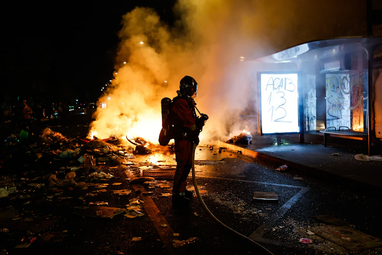 Un bombero en la calle por la noche ante un fuego ocasionado por los disturbios en Francia junto a una marquesina de autobús
