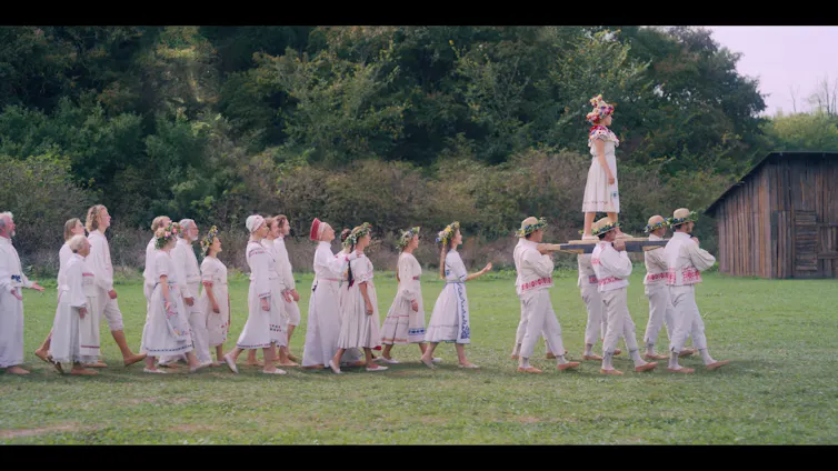 Procession of people dressed in white, with the shape of a face in the woodsy background
