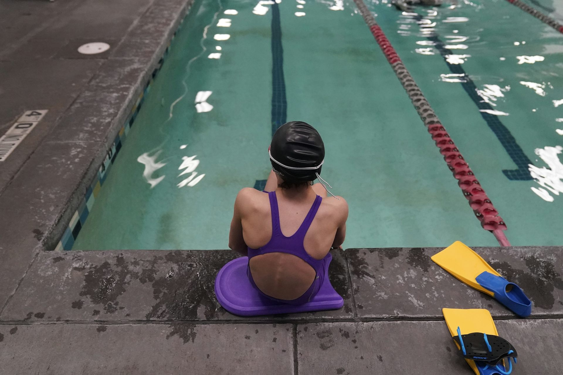 A child in a purple swimsuit, seen from behind, sits at the edge of a pool beside yellow swim fins.
