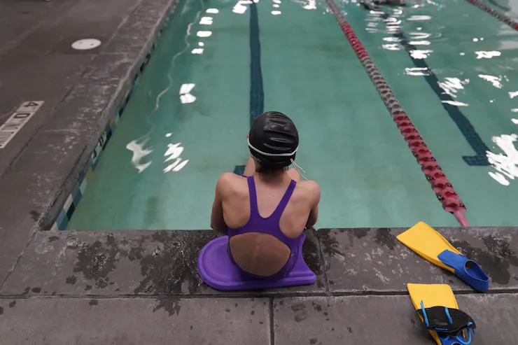 A child in a purple swimsuit, seen from behind, sits at the edge of a pool beside yellow swim fins.