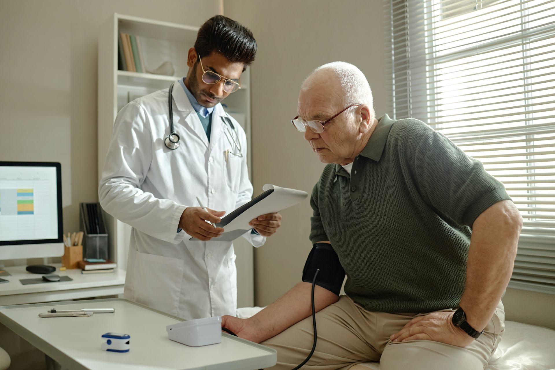An older man has his blood pressure taken by a young male doctor who is wearing a white lab coat.