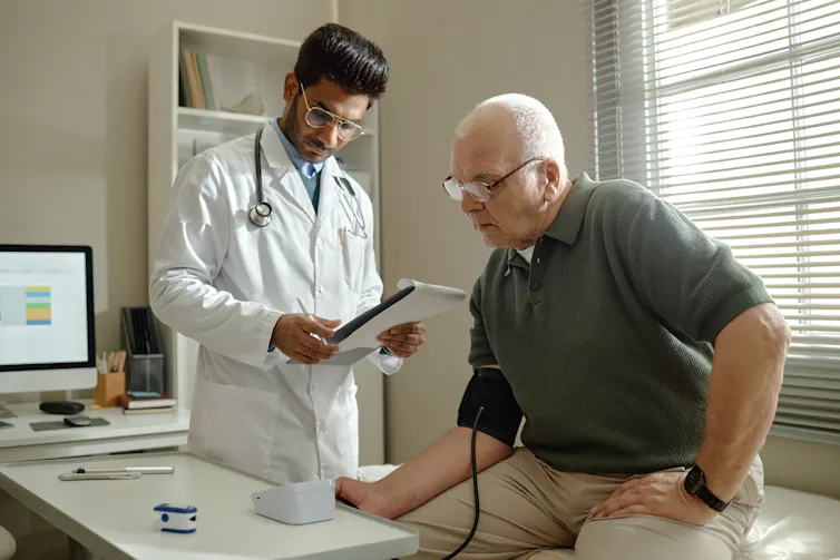 An older man has his blood pressure taken by a young male doctor who is wearing a white lab coat.