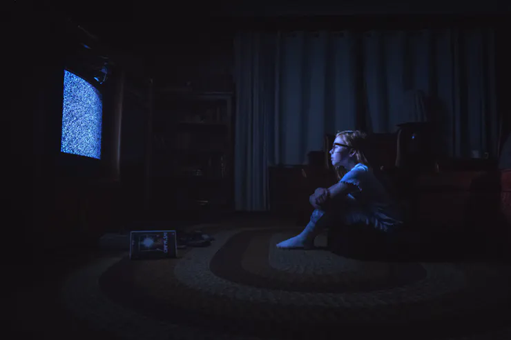 Film still of a young girl in a dark room, sitting before a TV filled with static
