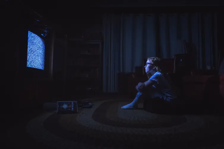 Film still of a young girl in a dark room, sitting before a TV filled with static