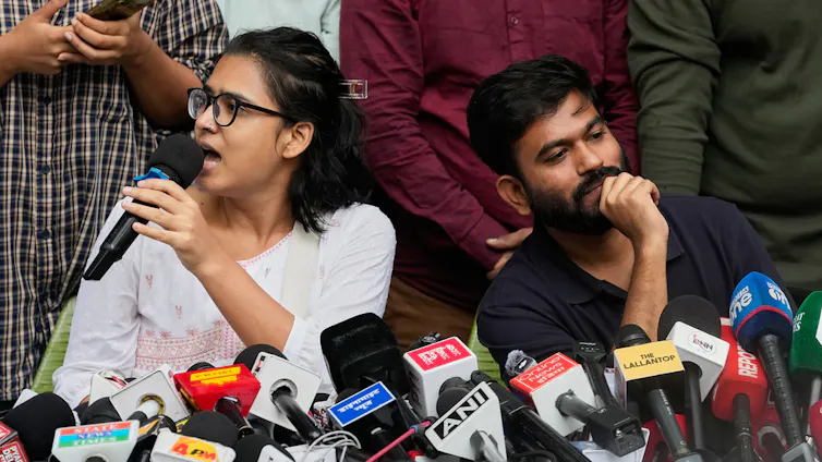 A man and a woman address an audience in front of microphones.