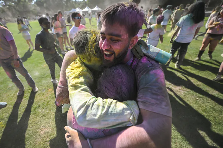 Three young people covered in different bright colors on their faces, hair and bodies hug in a field surrounded by other people.