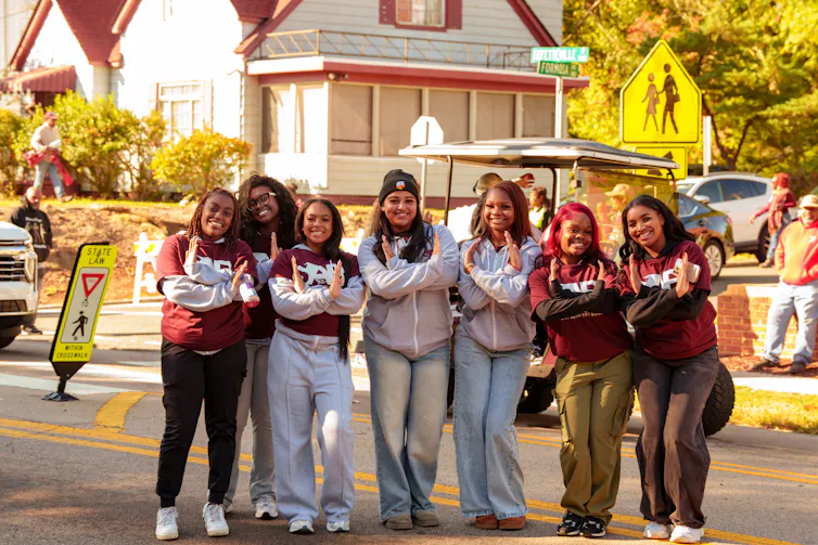 A group of young women wearing red and grey tops smile and cross their arms in a playful manner, as they stand close together on a residential street with a house behind them.
