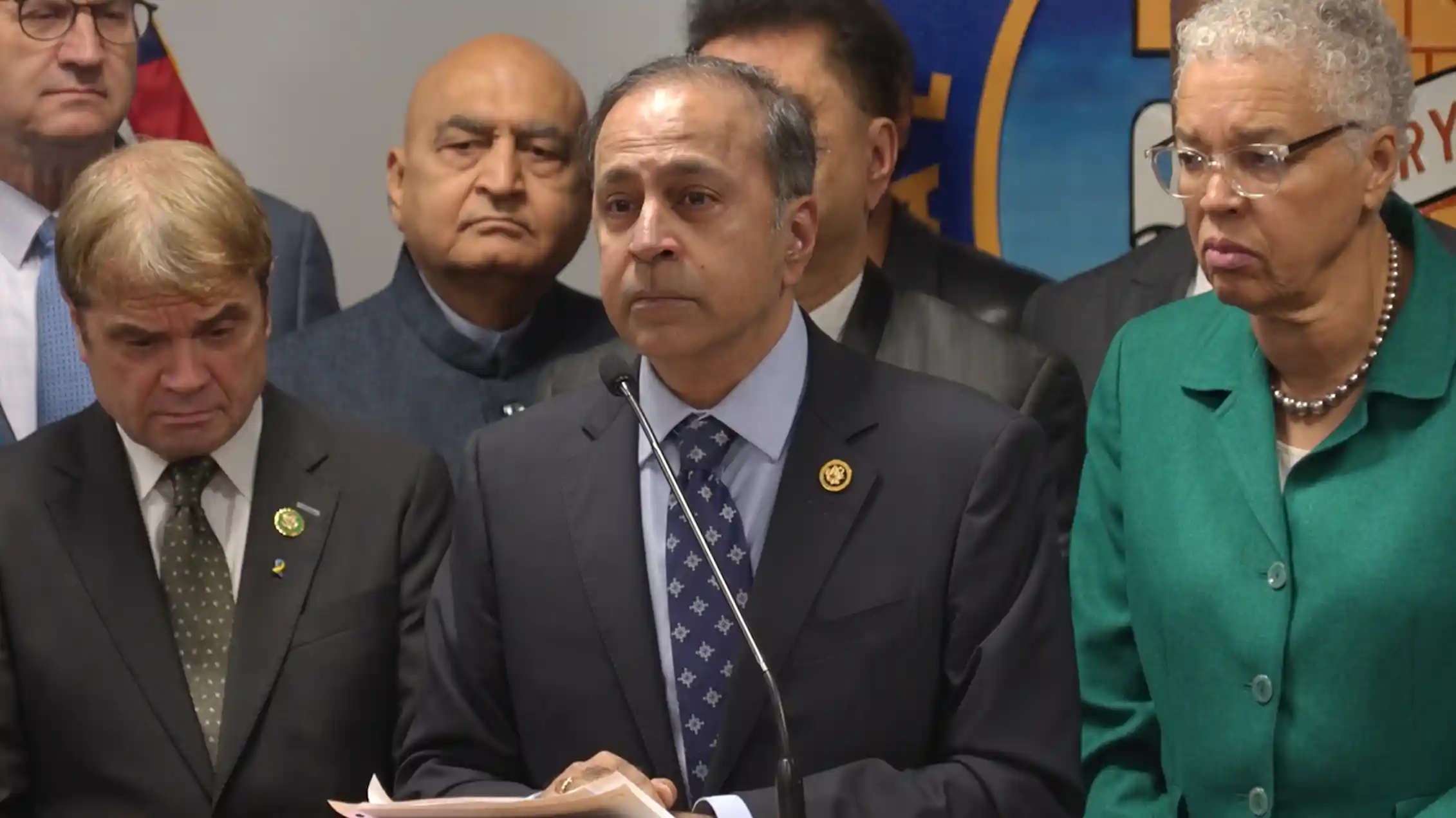 A group of men in suits stand behind a lectern, with one speaking into a microphone and a woman in a green dress beside them.
