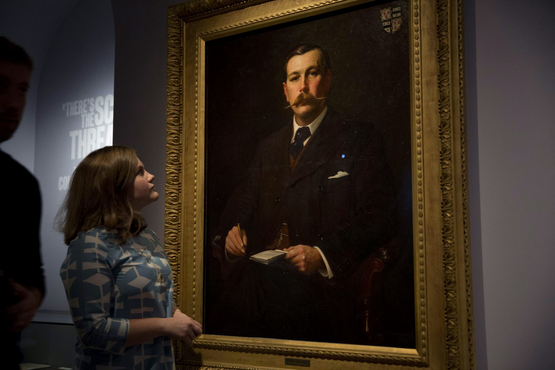 A woman looks up at the portrait of a man with a moustache.