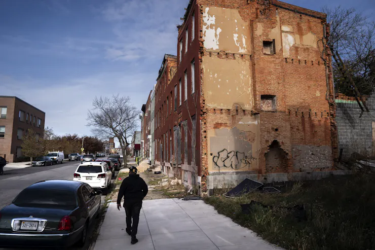 A man walks along a sidewalk past a row of vacant brick buildings.
