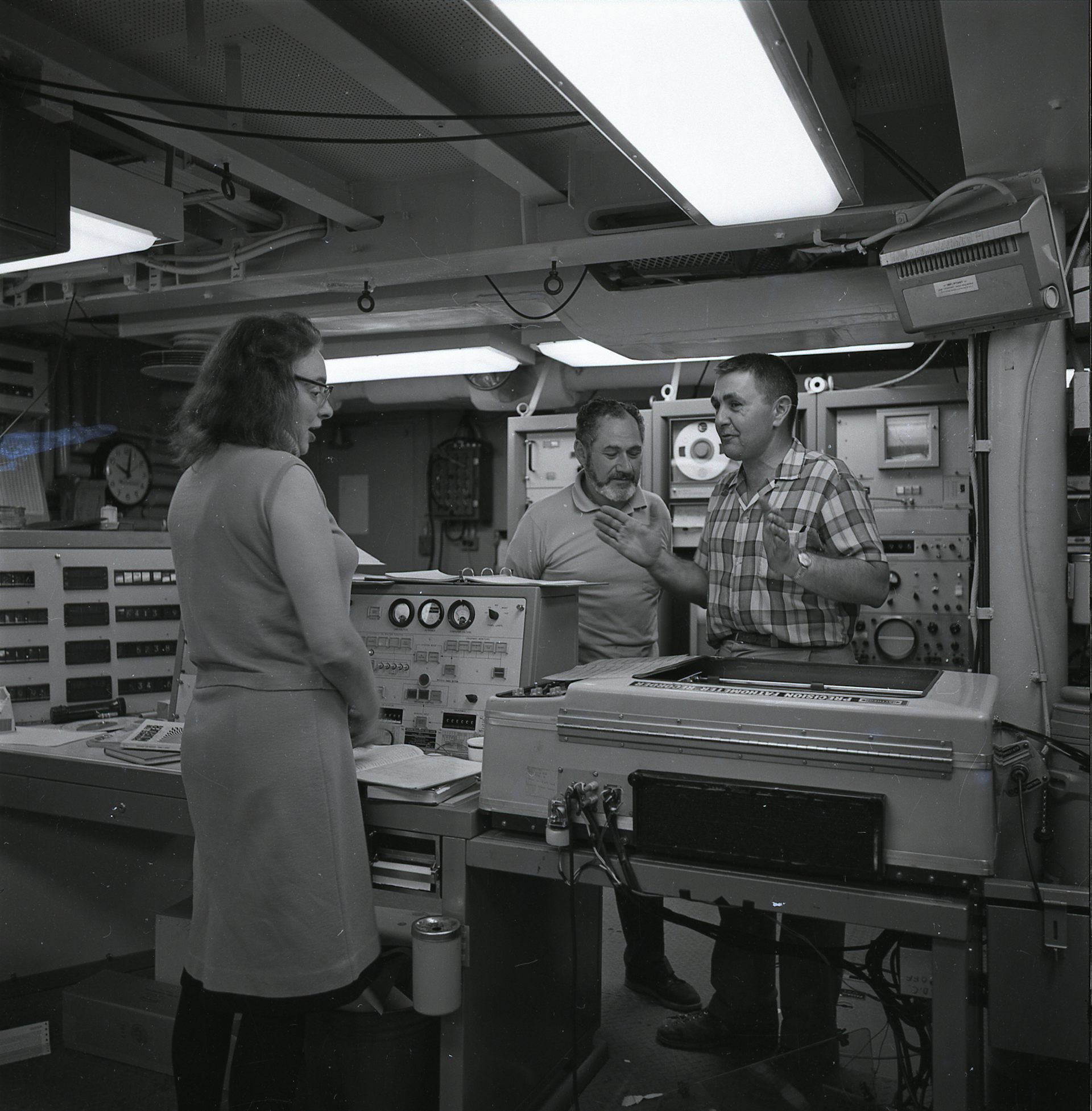 Black and white photo of Marie Tharp and two men talking surrounded by scientific equipment.