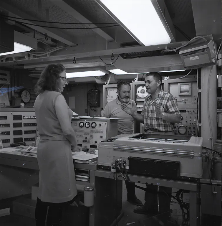 Black and white photo of Marie Tharp and two men talking surrounded by scientific equipment.