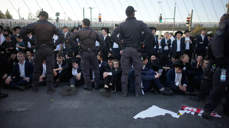 Three officers in gray uniforms stand with their backs toward the camera, near a crowd of men in suits and black hats in the middle of a road.