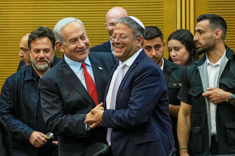 Two men in suits and ties grin and shake hands as several other people watch on.