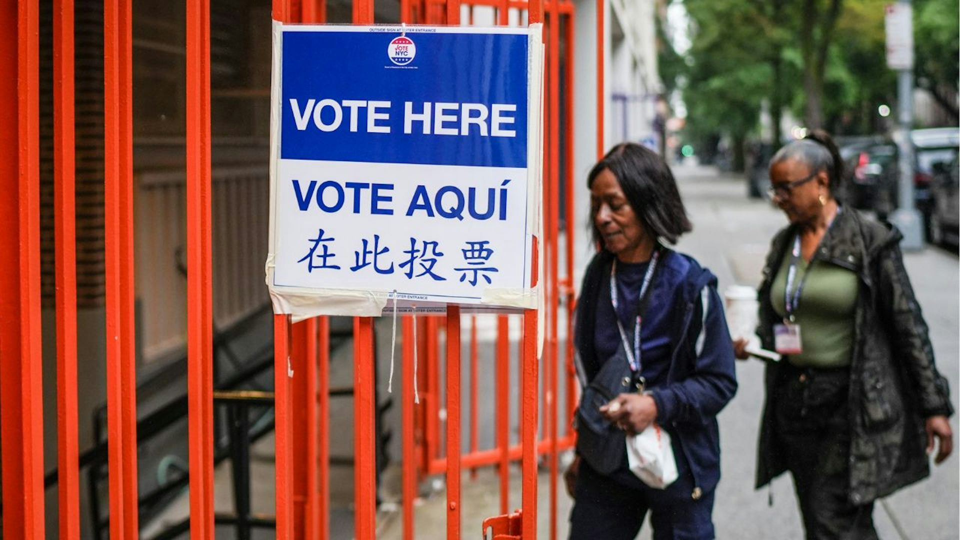 Two women approach a sign that says "vote here" in multiple languages. They are walking beside orange bars on a sidewalk.