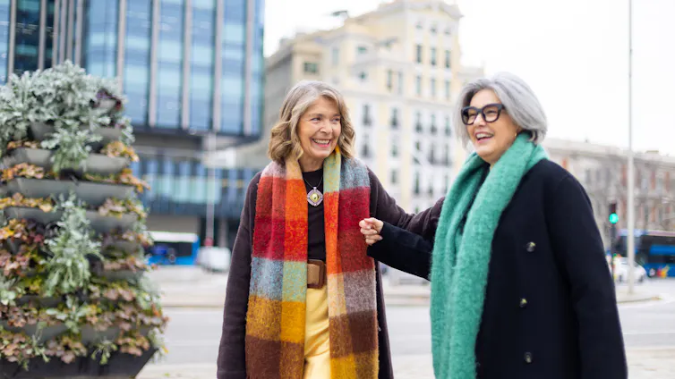 Two older women wearing scarves and winter coats laugh while they walk in a city.