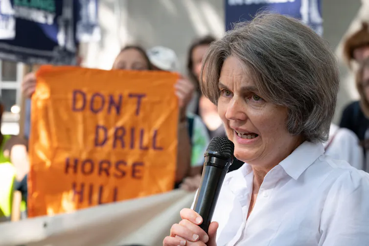 Woman with microphone, 'don't drill horse hill' sign in background