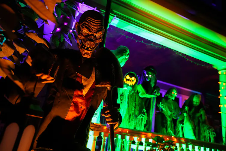 A ghoulish sculpture is illuminated orange in front of a porch with other Halloween decorations on it lit up with green lighting.