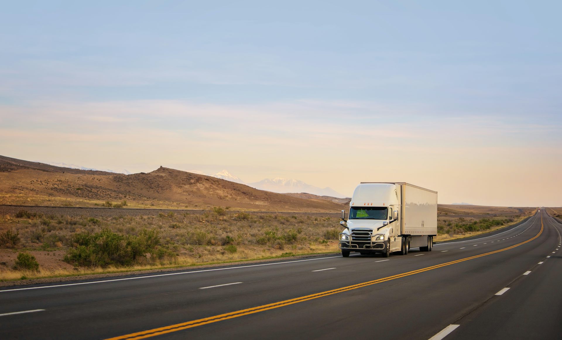 A white semi-truck pulling a white trailer drives down a highway, with hills in the background.